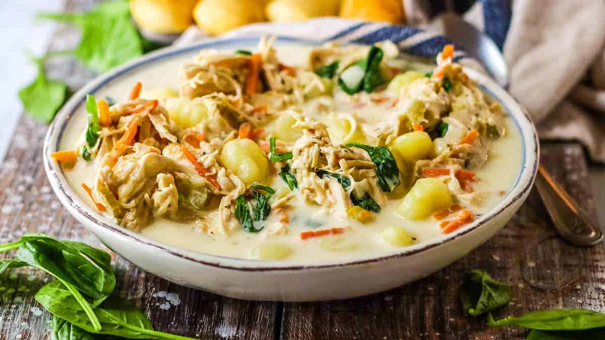 A bowl of creamy soup with shredded chicken, gnocchi, spinach, and carrots, served on a wooden table with fresh spinach leaves nearby.