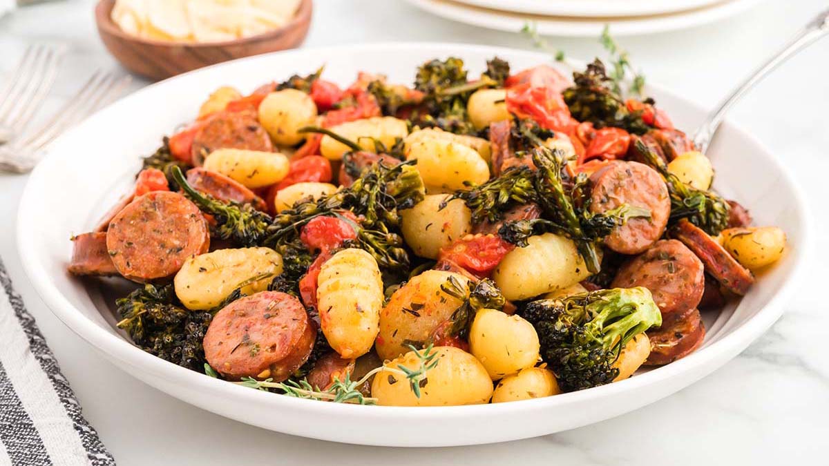 A white plate filled with gnocchi, sliced sausage, roasted broccolini, and cherry tomatoes, garnished with herbs.