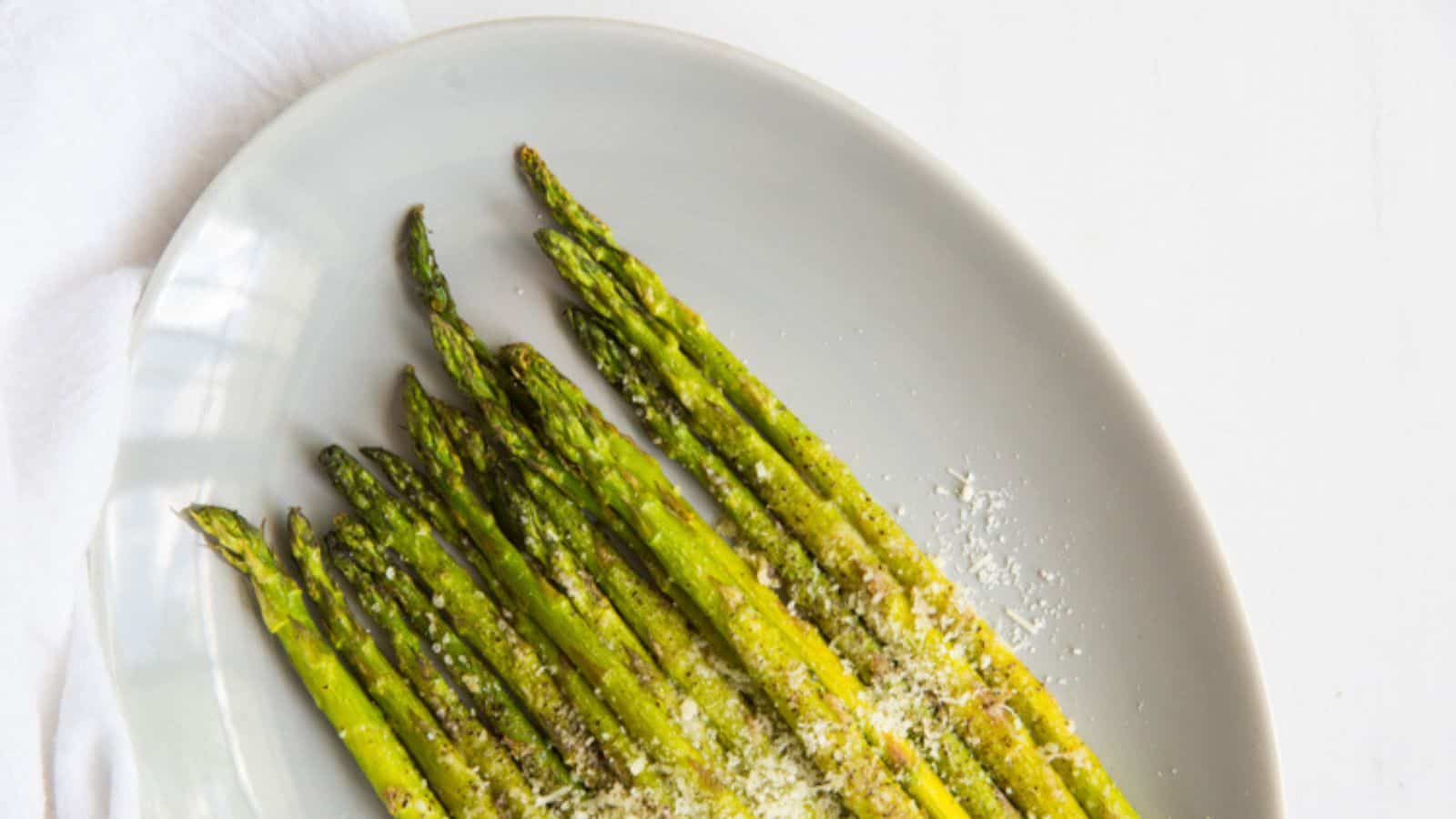 A white plate with cooked asparagus spears sprinkled with grated cheese, set on a white surface.