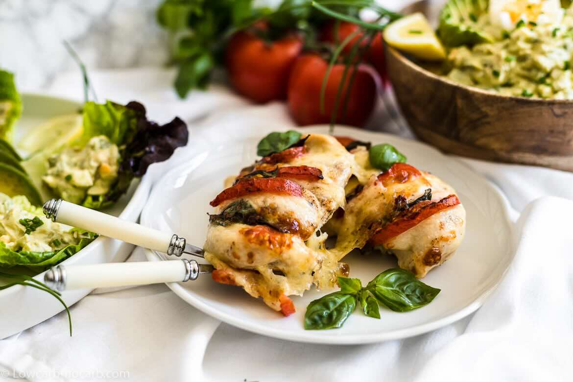 A plate with baked chicken breast stuffed with cheese, tomatoes, and herbs, garnished with basil, and placed next to two forks and a green salad.