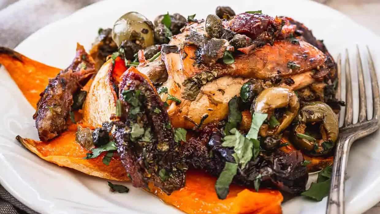 A plate of roasted chicken topped with herbs, olives, and capers, served alongside roasted sweet potato wedges. A fork is placed on the plate.