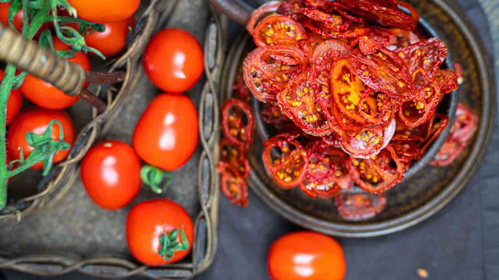 A basket of fresh tomatoes next to a plate of sliced sun-dried tomatoes on a dark surface.