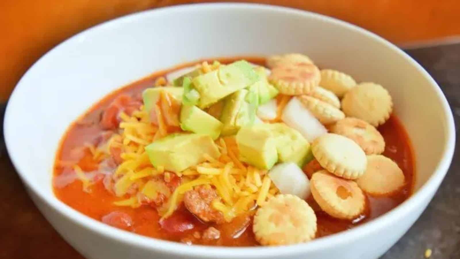 A bowl of chili topped with shredded cheddar cheese, diced avocado, chopped onion, and round oyster crackers.