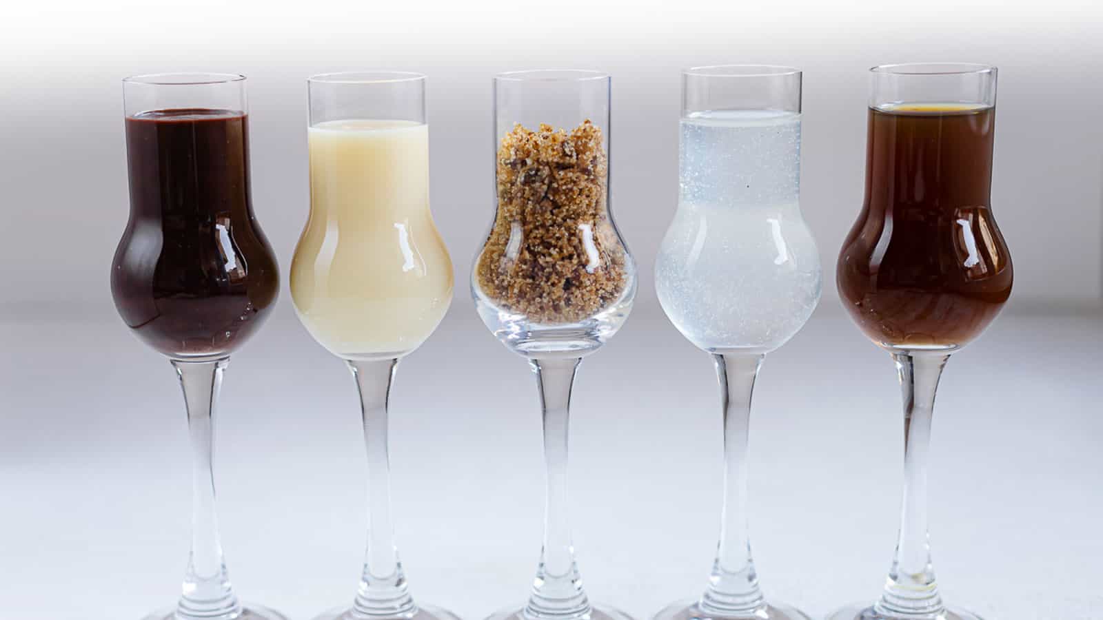 Five liqueur glasses are lined up, each containing a different substance: a dark liquid, a light creamy liquid, brown sugar crystals, a clear liquid, and a dark amber liquid.