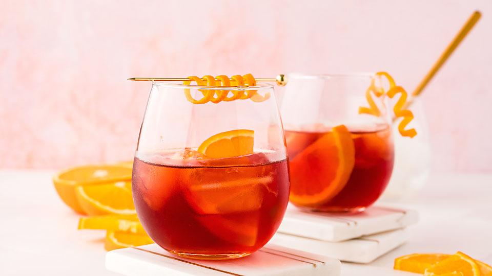 Two glasses of a red cocktail with ice and orange slices, garnished with orange twists, sit on white coasters with more orange slices in the background.