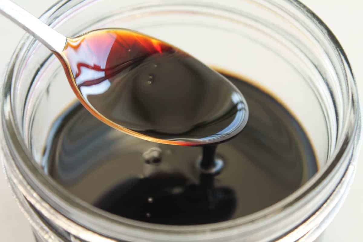 A spoonful of dark liquid, likely soy sauce or molasses, held over an open glass jar filled with the same liquid.