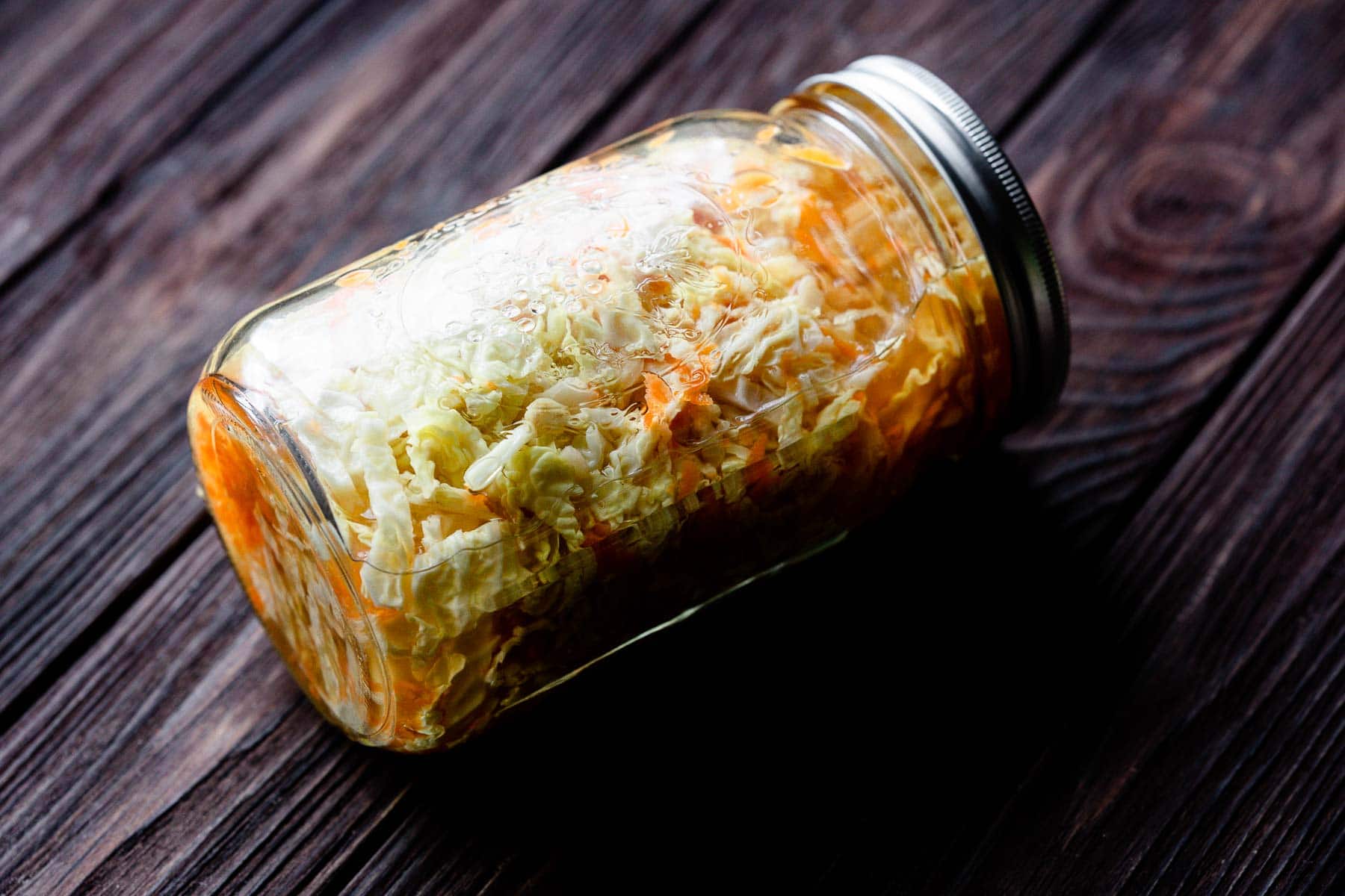 A glass jar filled with shredded cabbage and carrots sits on its side on a dark wooden surface.