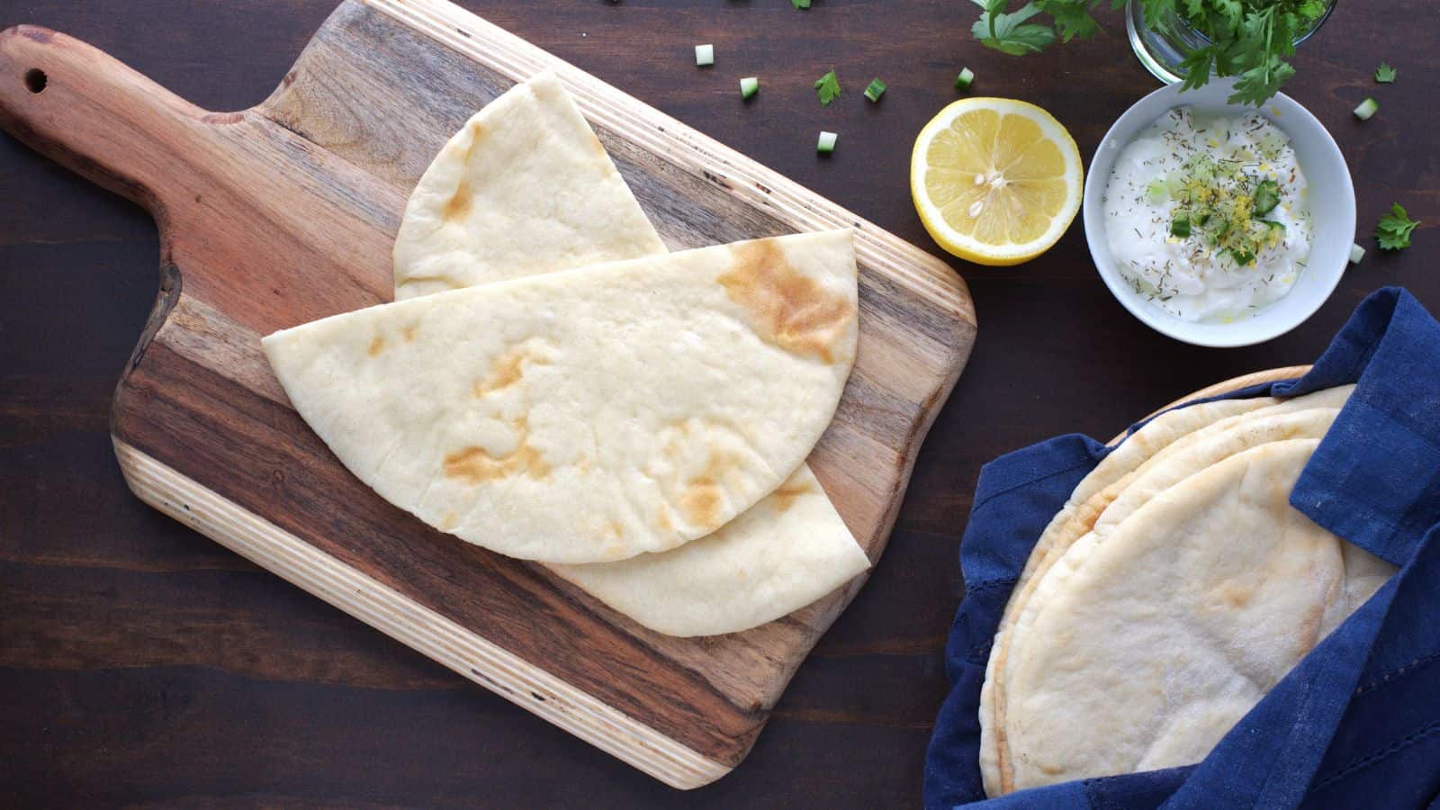 Two pieces of pita bread on a wooden cutting board, with a bowl of dip, lemon half, fresh herbs, and more pita bread in a cloth-lined basket nearby.
