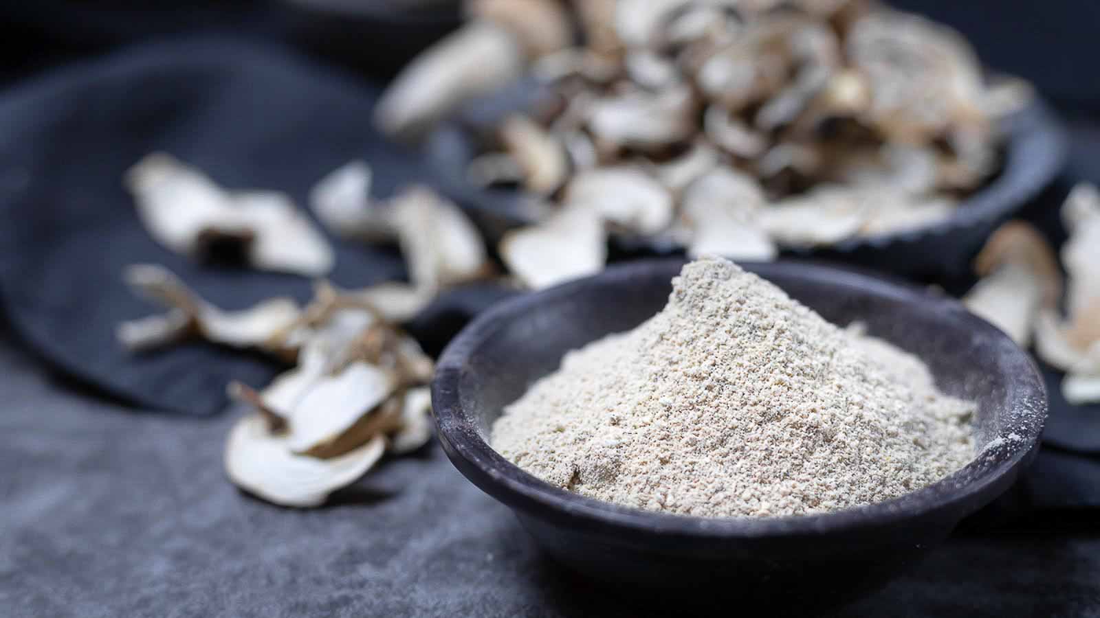 A small black bowl filled with mushroom powder sits in front of scattered dried mushroom slices on a dark surface.
