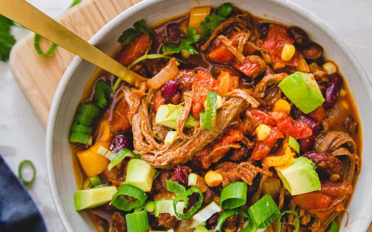 A bowl of chili with shredded meat, kidney beans, corn, diced tomatoes, avocado, and sliced green onions, served with a gold spoon.