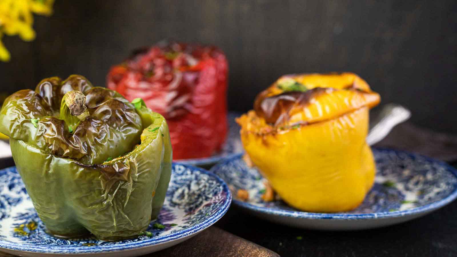 Three roasted stuffed bell peppers—green, red, and yellow—are served on separate blue patterned plates against a dark background.