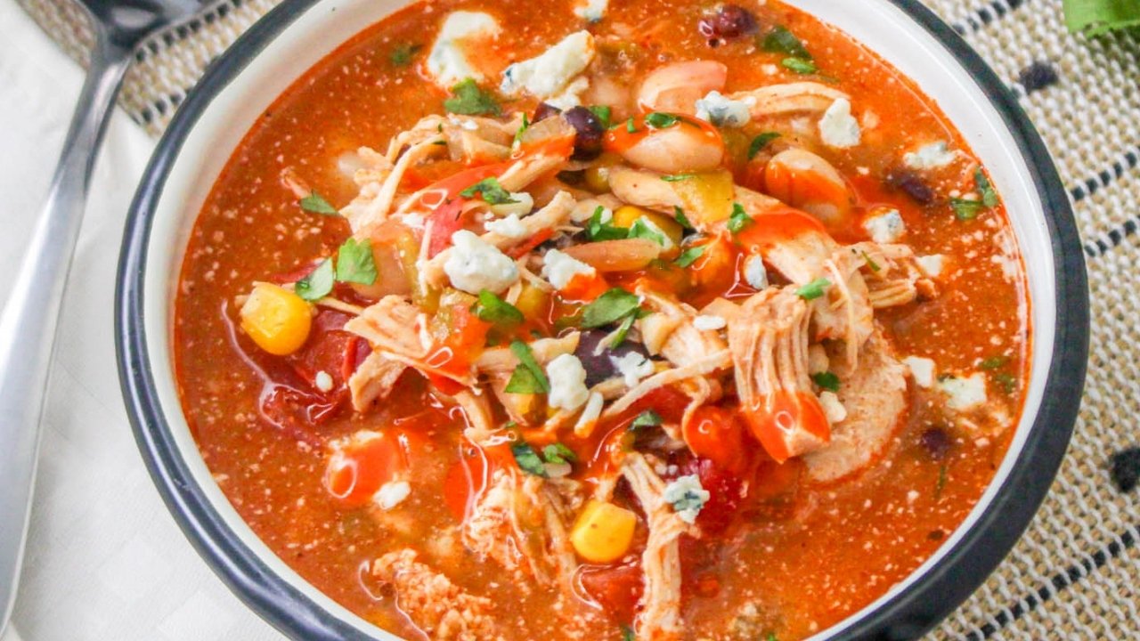 A bowl of chicken chili soup with beans, corn, tomatoes, shredded chicken, and crumbled cheese, garnished with chopped herbs.