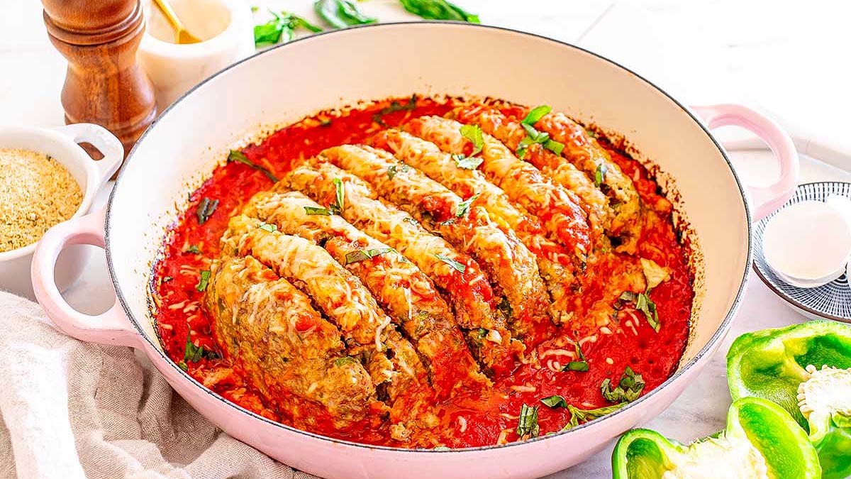 A round pan filled with sliced stuffed cabbage rolls in tomato sauce, garnished with herbs. Green bell peppers and ingredients are visible nearby.