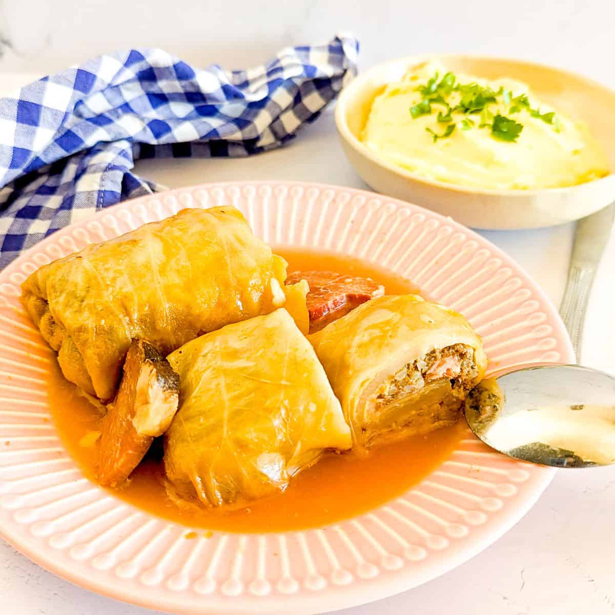 Three cabbage rolls in tomato sauce on a pink plate with a side bowl of mashed potatoes garnished with herbs; a spoon and blue checkered napkin nearby.