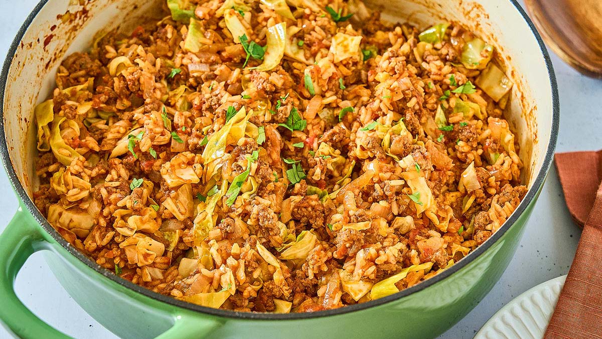 A green Dutch oven filled with cooked ground meat, rice, chopped cabbage, tomatoes, and herbs.