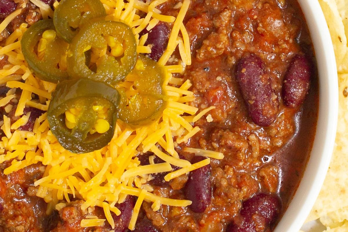 A bowl of chili with ground beef, kidney beans, shredded cheddar cheese, and sliced jalapeรฑos, served with tortilla chips on the side.