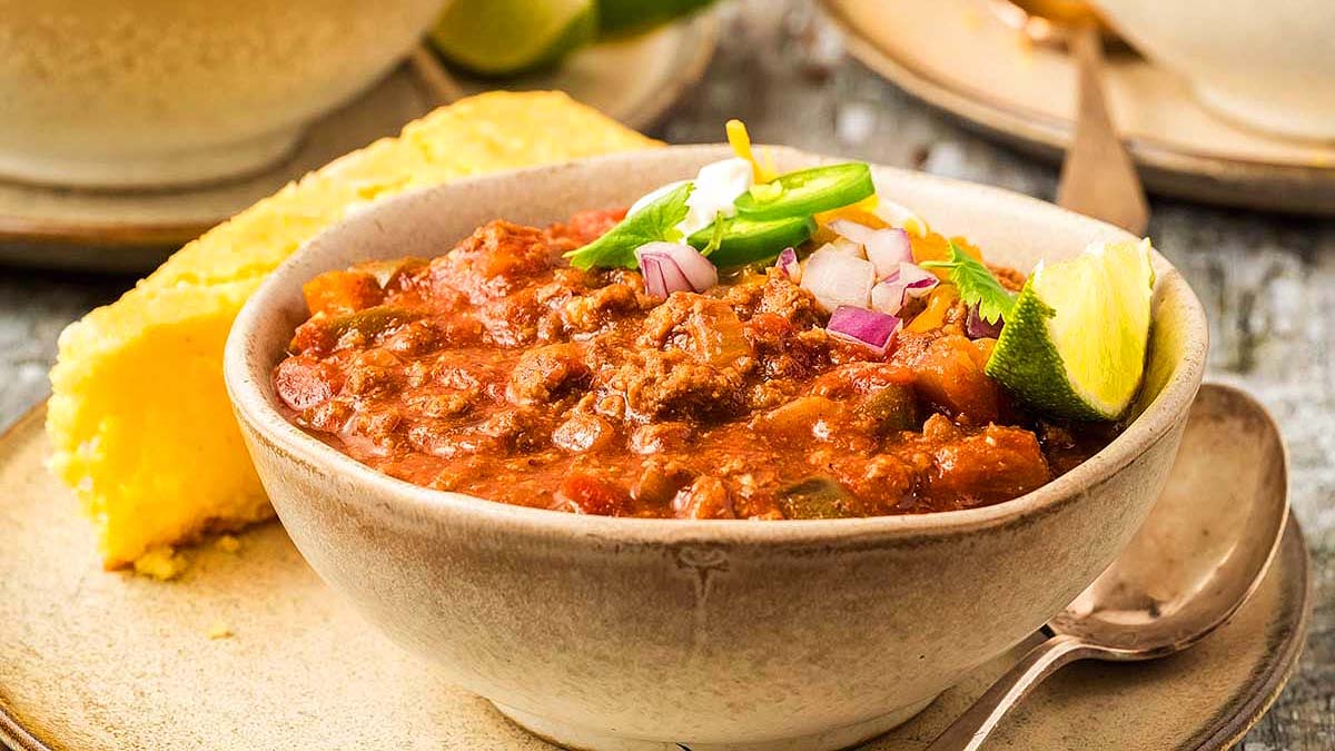 A bowl of chili topped with chopped onions, cheese, and green onions, served with a wedge of lime and a slice of cornbread on the side.
