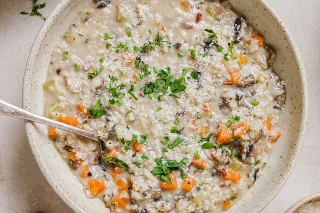 A bowl of creamy soup with visible chunks of carrots, mushrooms, grains, and herbs, garnished with chopped parsley.