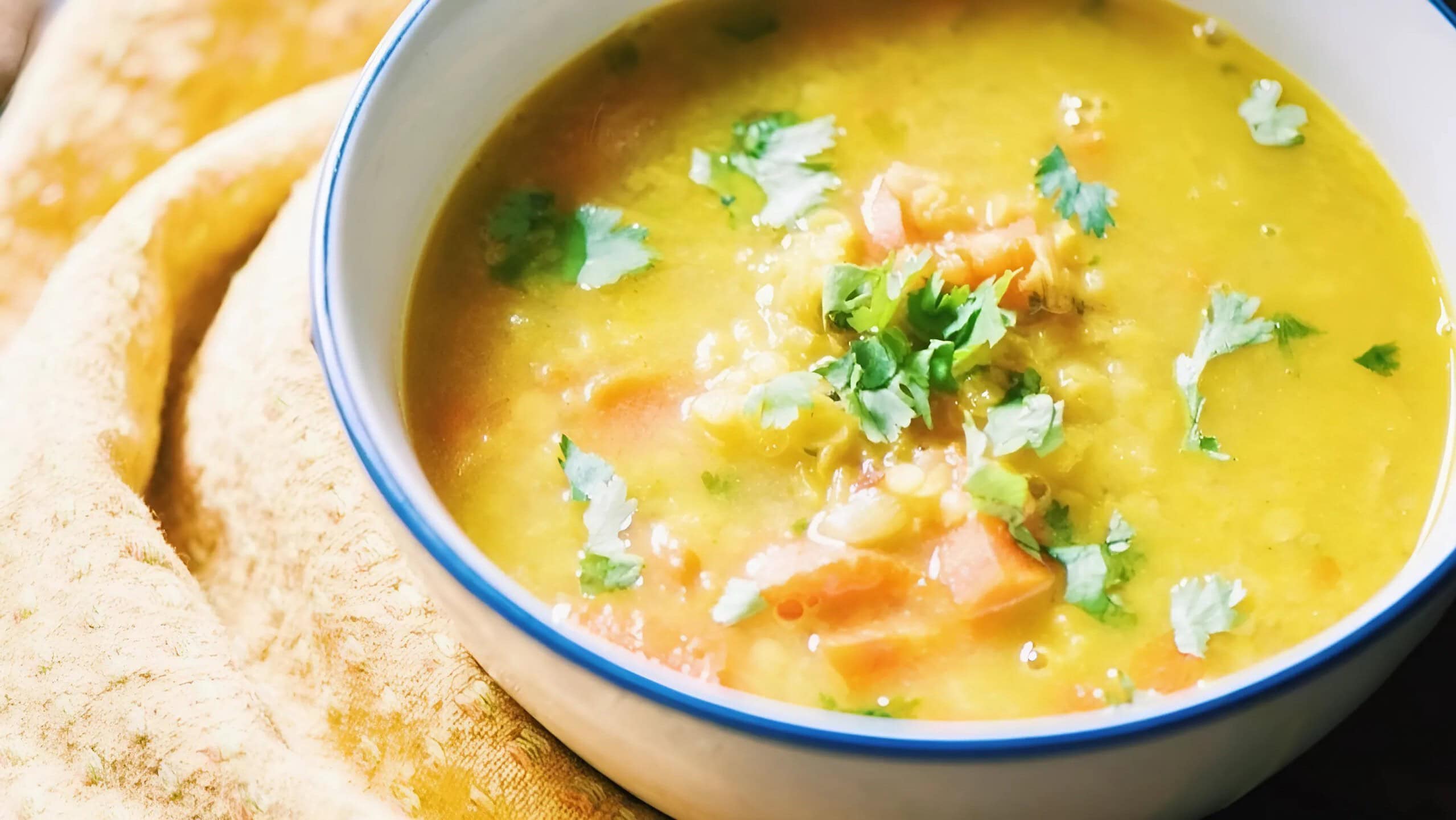 A close-up of a bowl of yellow lentil soup garnished with chopped cilantro and diced vegetables, placed on a yellow cloth.