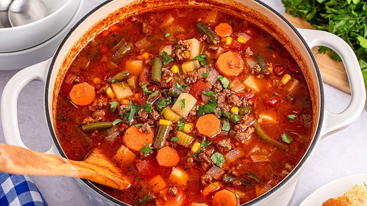 A pot of vegetable beef soup with chunks of carrots, potatoes, corn, green beans, and ground beef in a tomato broth, garnished with fresh herbs.