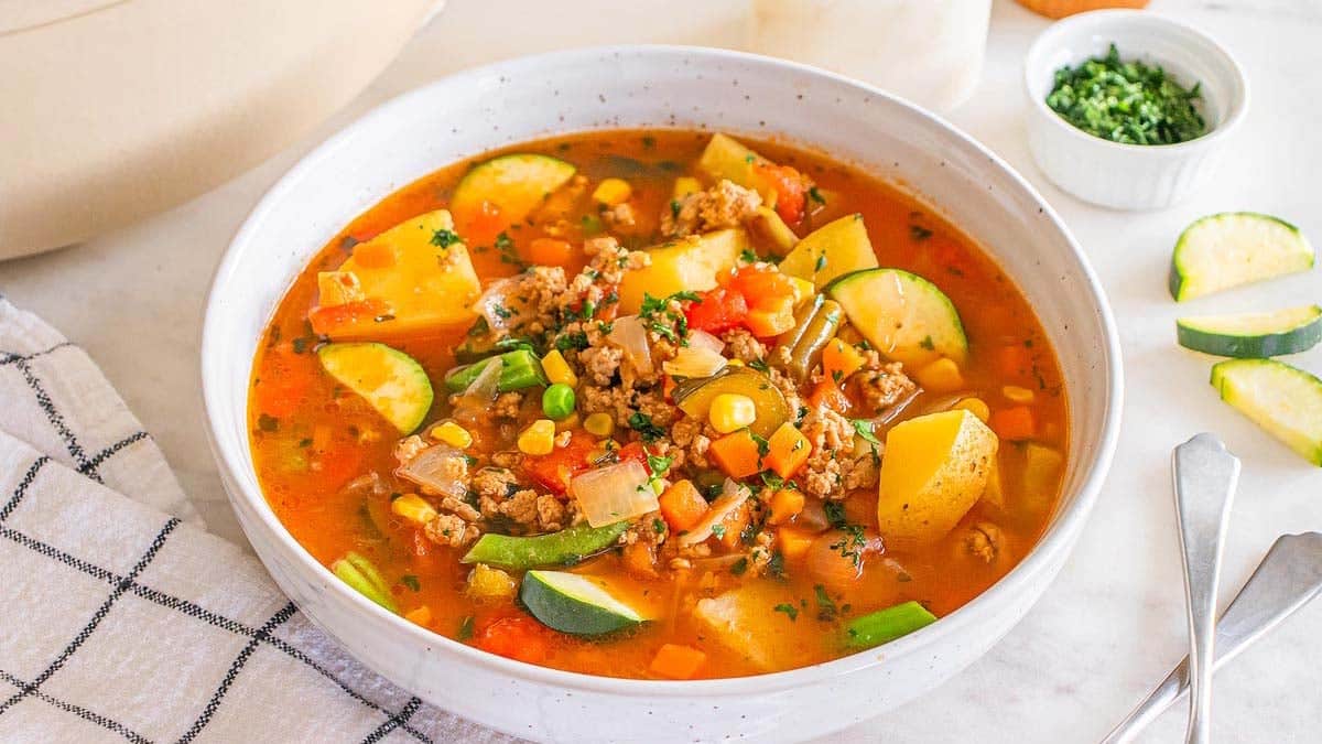 A bowl of chunky vegetable and ground meat soup with potatoes, zucchini, corn, peas, and carrots on a white table with a napkin and a small bowl of herbs.