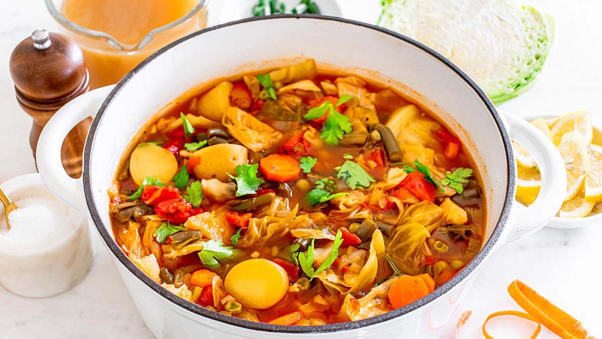 A white pot filled with vegetable soup containing potatoes, carrots, cabbage, tomatoes, and herbs sits on a countertop surrounded by soup ingredients and seasonings.