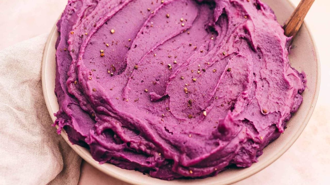 A bowl of mashed purple sweet potatoes topped with cracked black pepper, with a wooden spoon resting on the side.