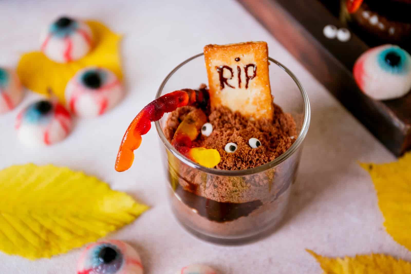 A glass cup filled with chocolate dessert, candy eyes, a gummy worm, and a cookie marked RIP, surrounded by fake eyeballs and yellow leaves.