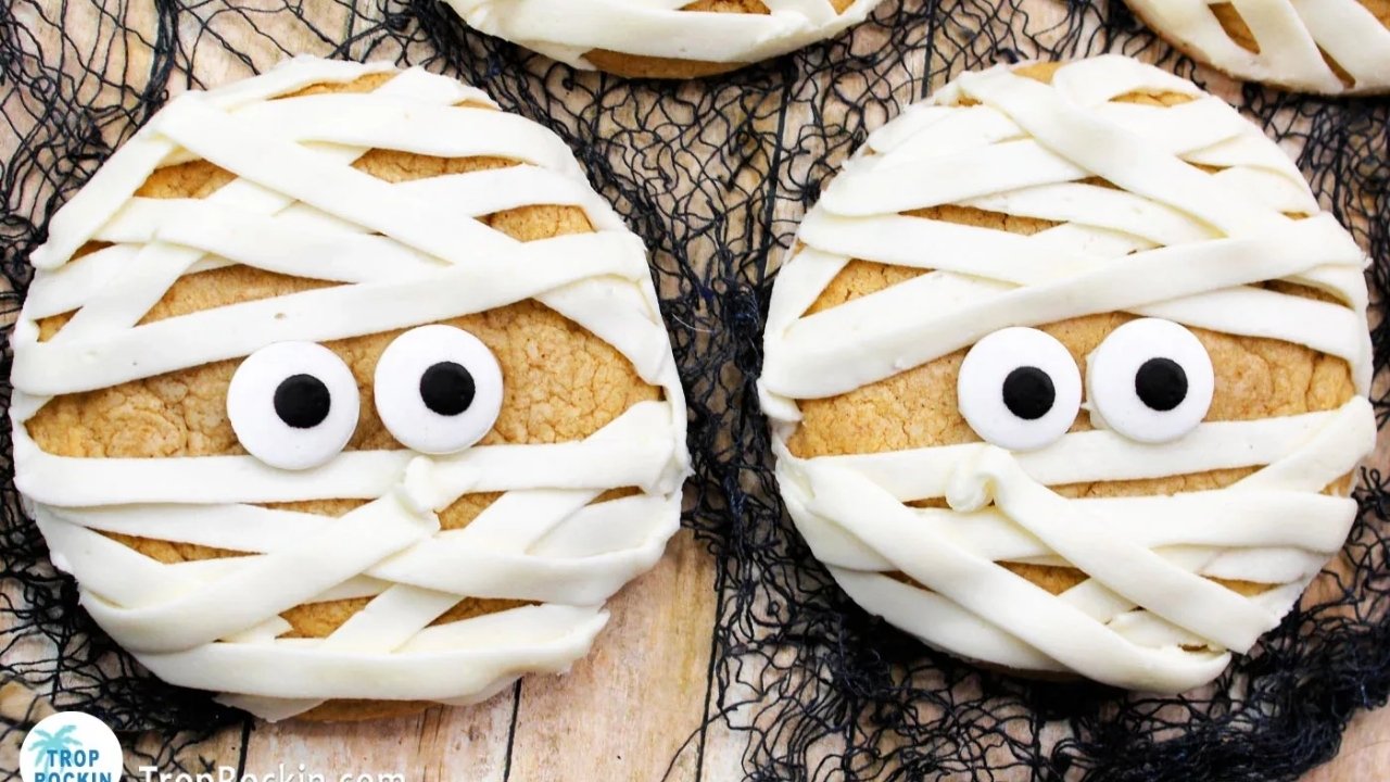 Two round cookies decorated with white icing to look like mummy faces, each with two round candy eyes, placed on a wooden surface.