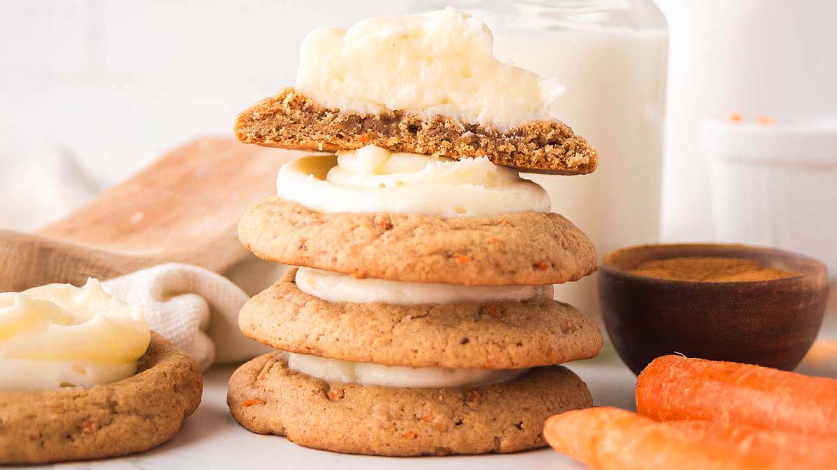 A stack of three frosted cookies sits on a white surface with a halved frosted cookie on top; carrots and a glass of milk are in the background.