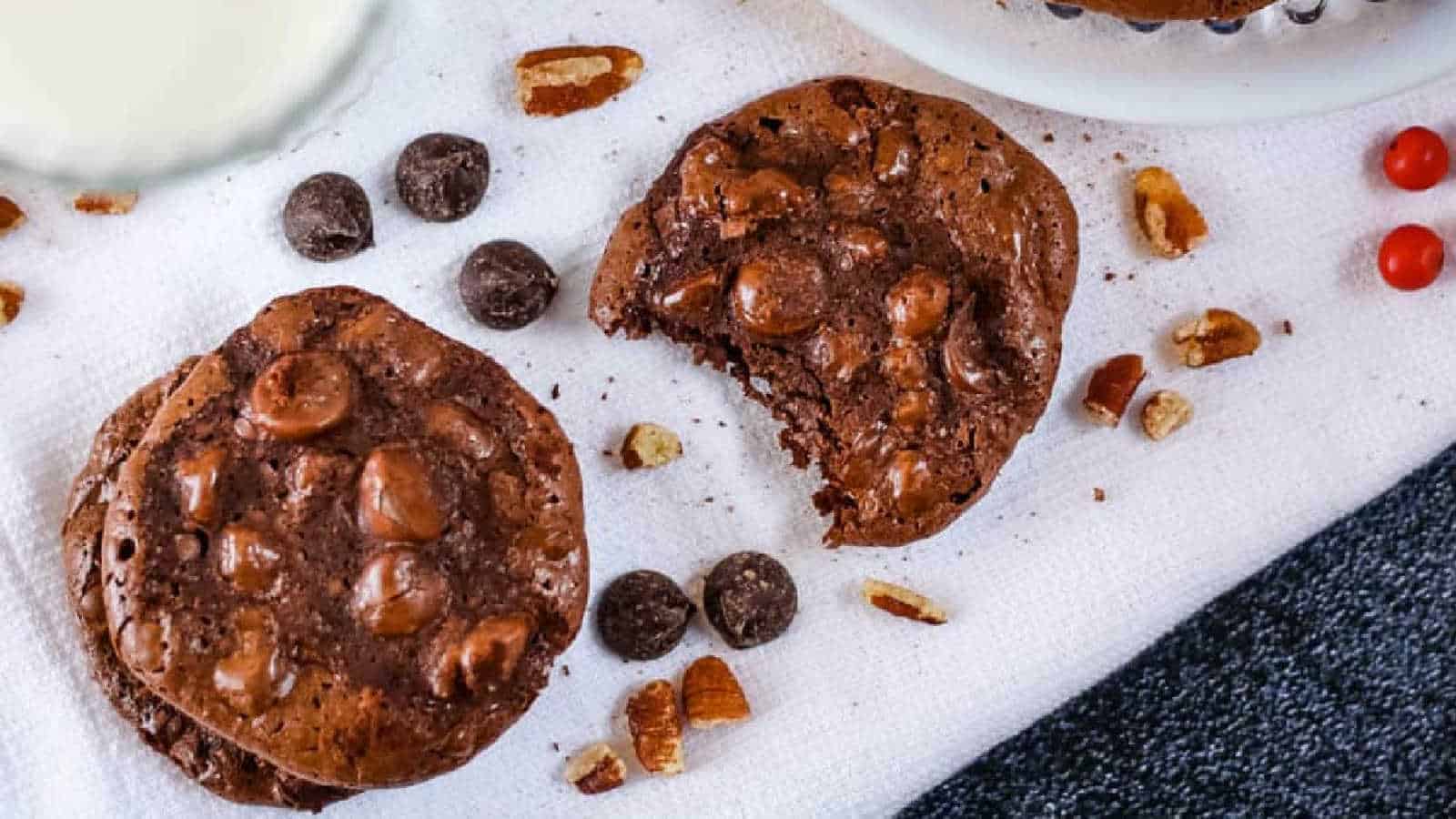 Three chocolate cookies with chocolate chips and nuts are on a white cloth, with one cookie partially eaten. Scattered chocolate chips, nuts, and a glass of milk are also visible.