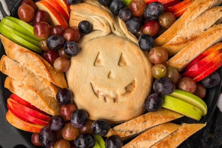 A round baked pastry with a jack-o’-lantern face is surrounded by sliced apples, grapes, and baguette slices on a black platter.