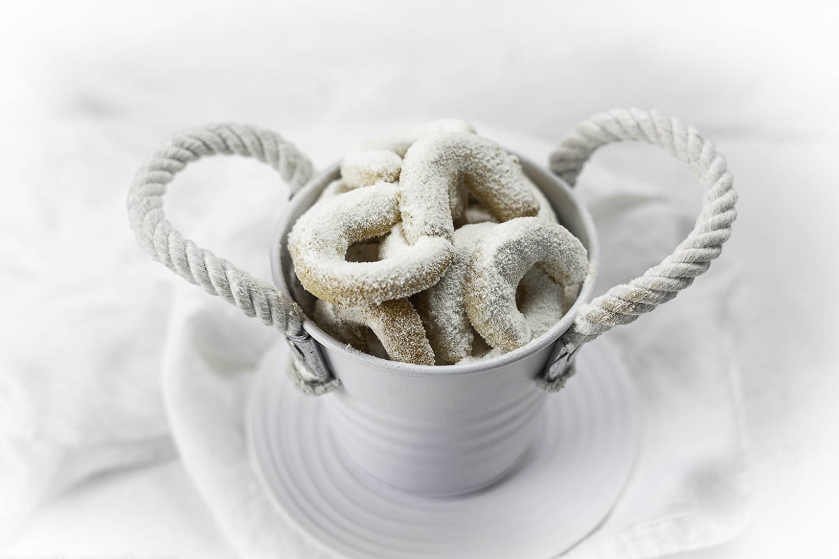 A small white bucket with rope handles contains powdered sugar-covered ring cookies, placed on a white plate and white cloth background.