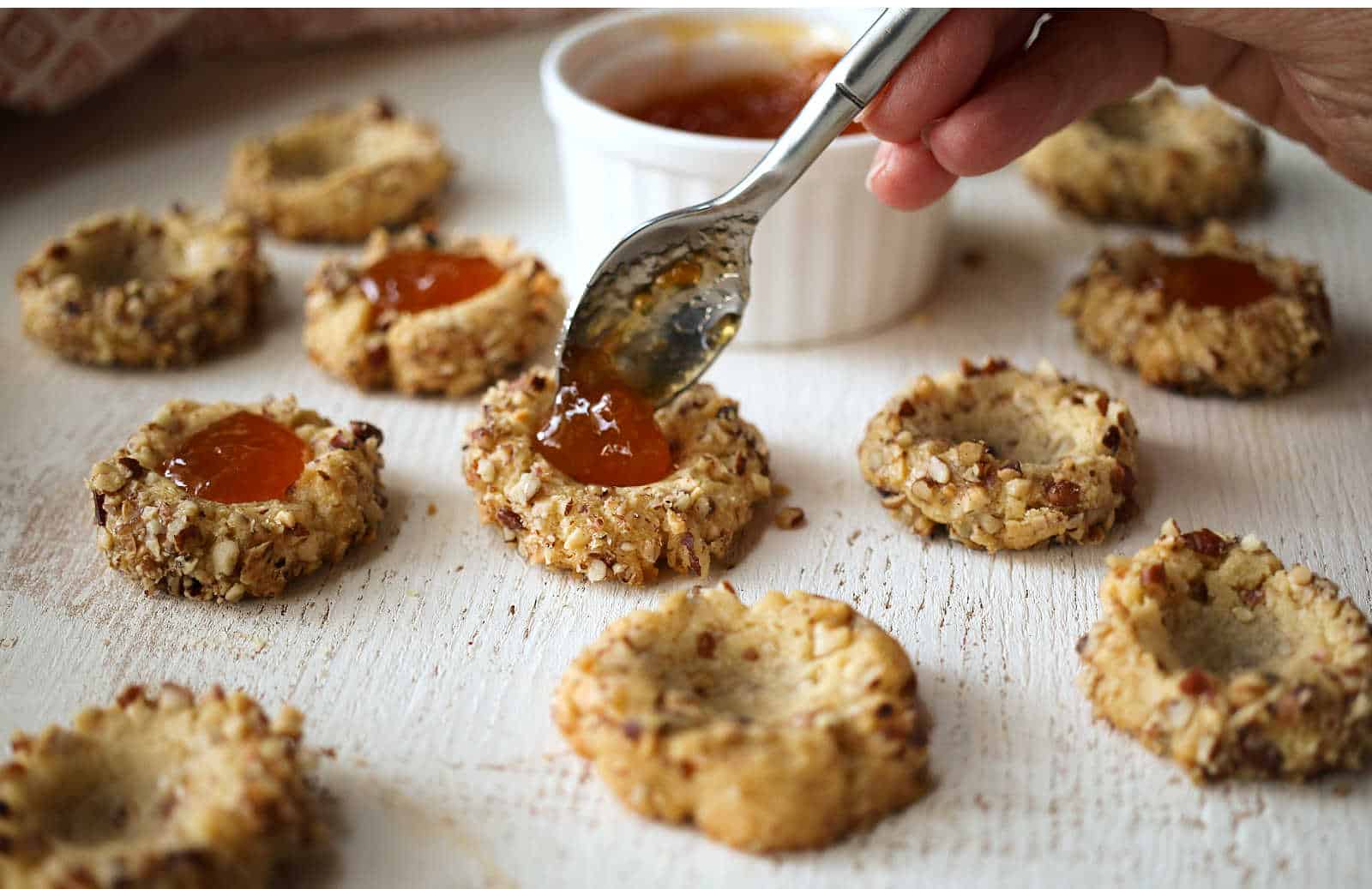 A hand uses a spoon to fill nut-coated thumbprint cookies with orange jam; several cookies and a ramekin of jam are on a white surface.
