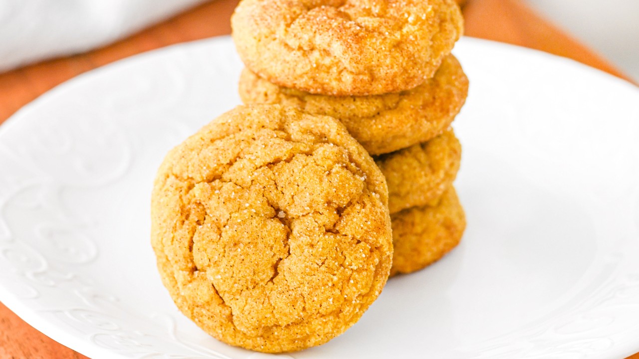 A stack of four golden brown cookies on a white plate, with one cookie leaning against the stack.