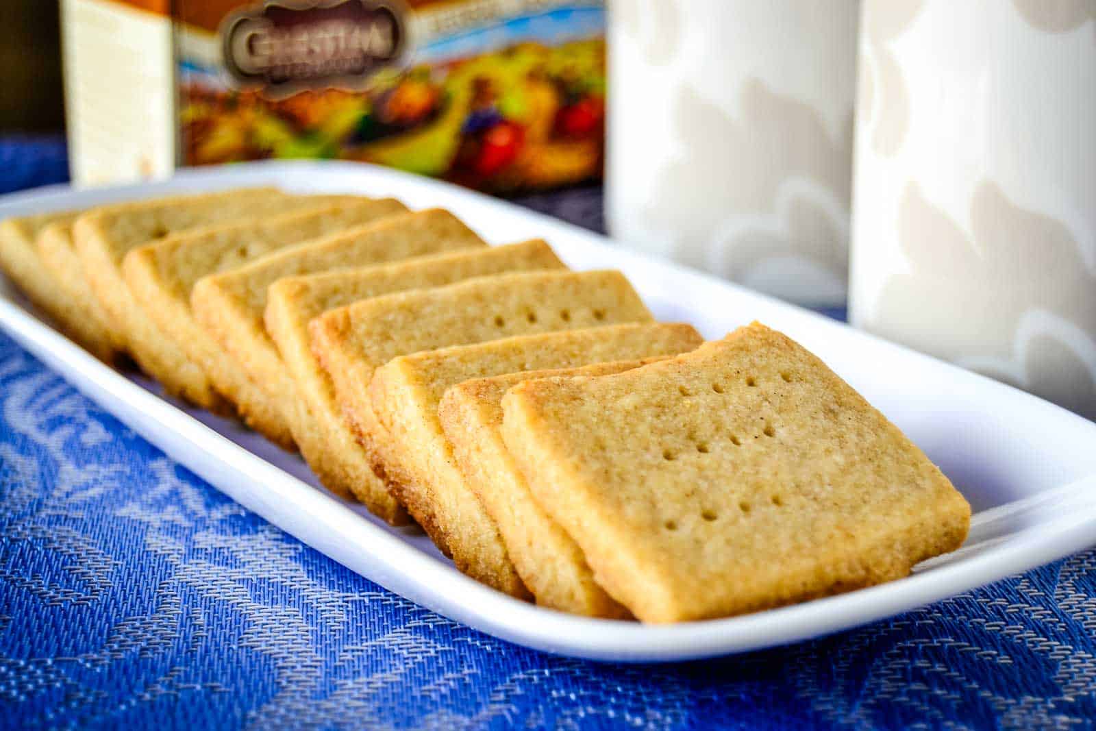 A white rectangular plate with rows of square shortbread cookies on a blue textured tablecloth. A box and two cylindrical containers are visible in the background.