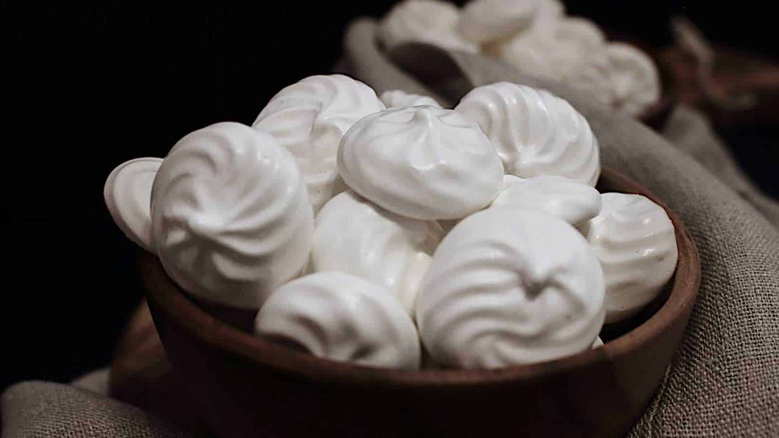 A wooden bowl filled with small, white meringue cookies, with additional meringues scattered around on a dark background.