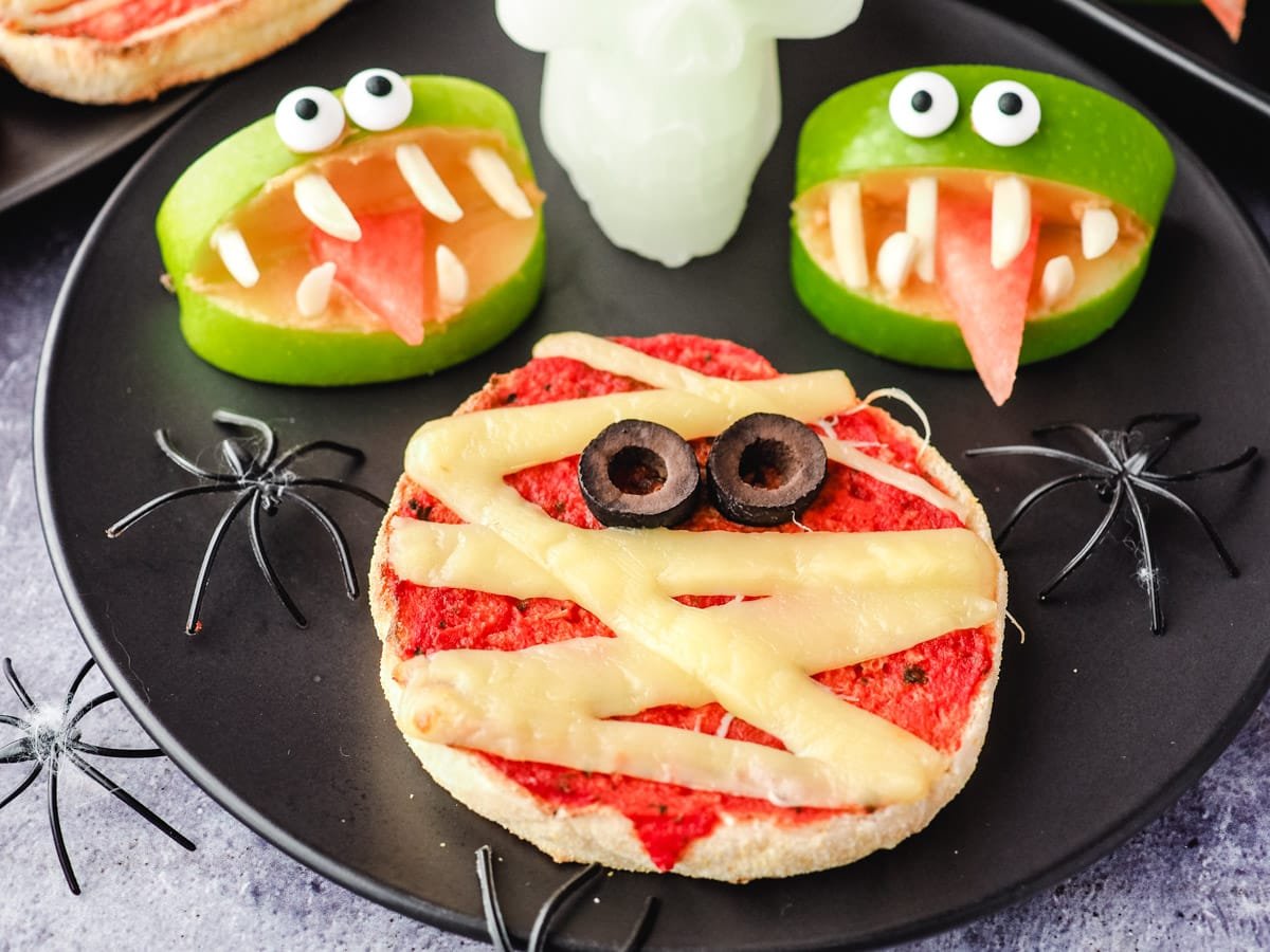 A mini pizza decorated as a mummy with cheese and olives, next to two green apple slices made to look like monsters, and plastic spiders on a black plate.
