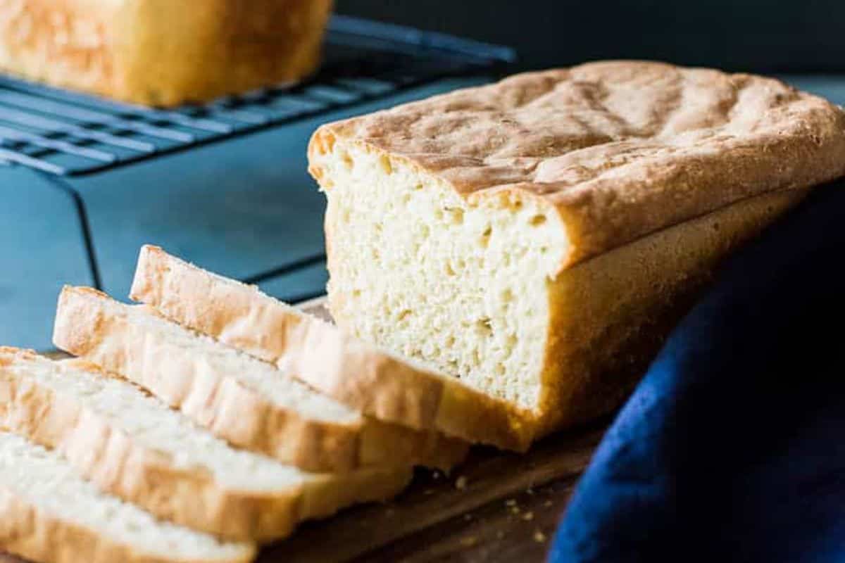 A loaf of bread with several slices cut, resting on a wooden board, with another loaf cooling on a wire rack in the background.
