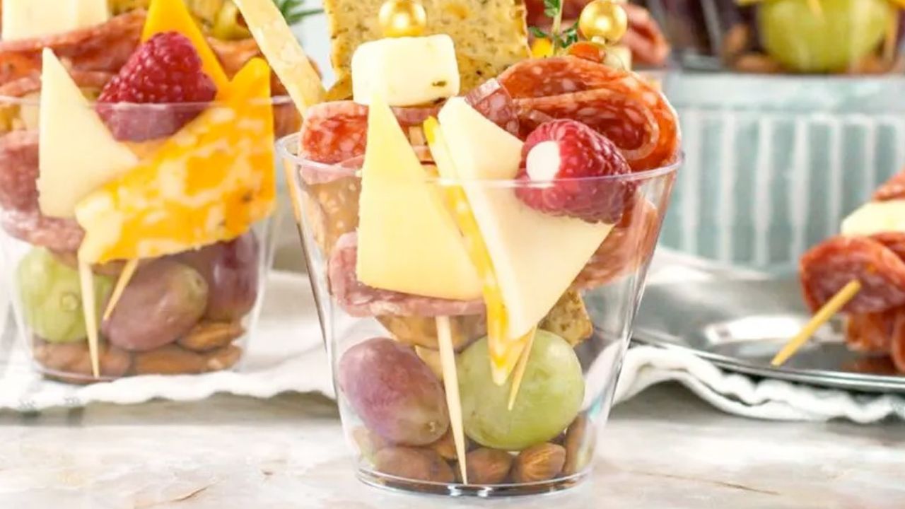 A clear cup filled with grapes, almonds, cubed and sliced cheese, salami, crackers, and raspberries, arranged on skewers.