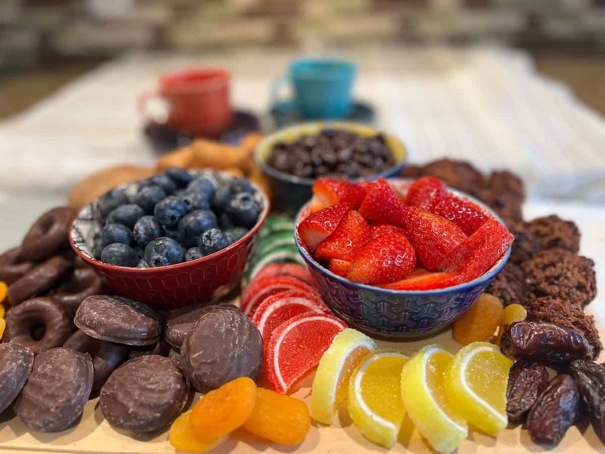 A platter with bowls of blueberries and strawberries, assorted dried fruits, chocolate-covered snacks, and coffee cups in the background on a striped cloth.