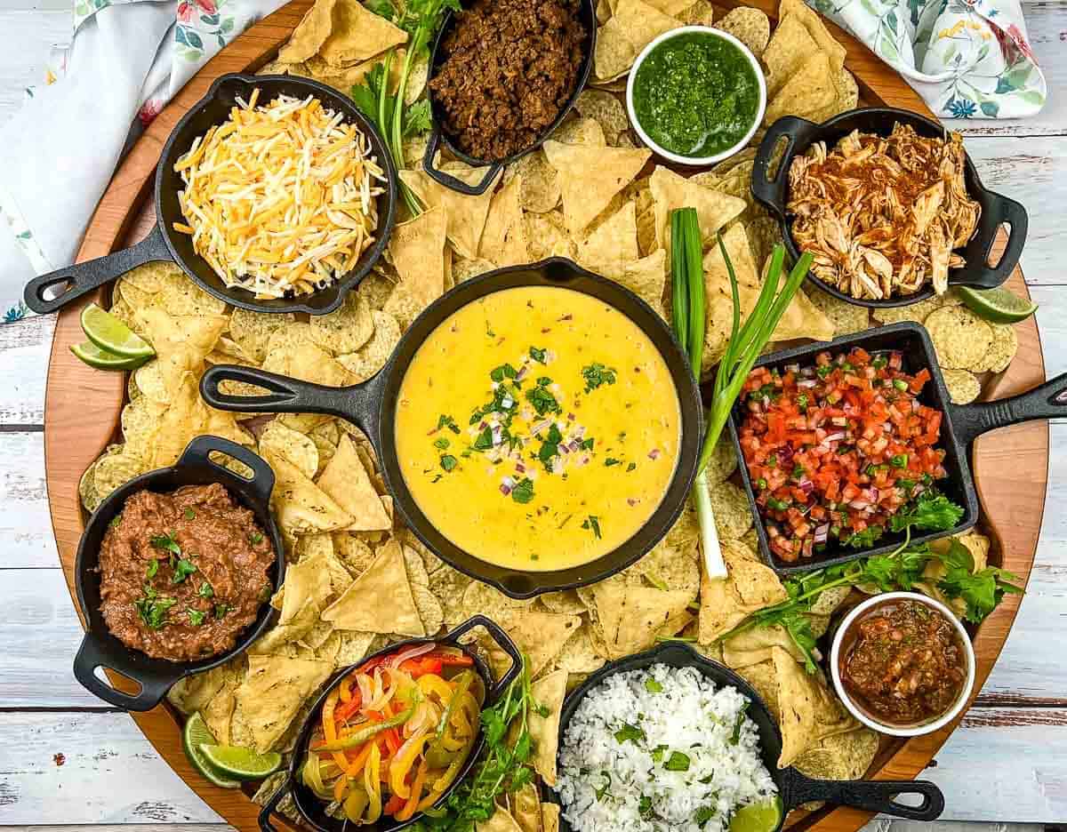 A platter with tortilla chips surrounded by bowls of queso, shredded cheese, ground beef, shredded chicken, refried beans, vegetables, rice, salsa, pico de gallo, and green sauce.