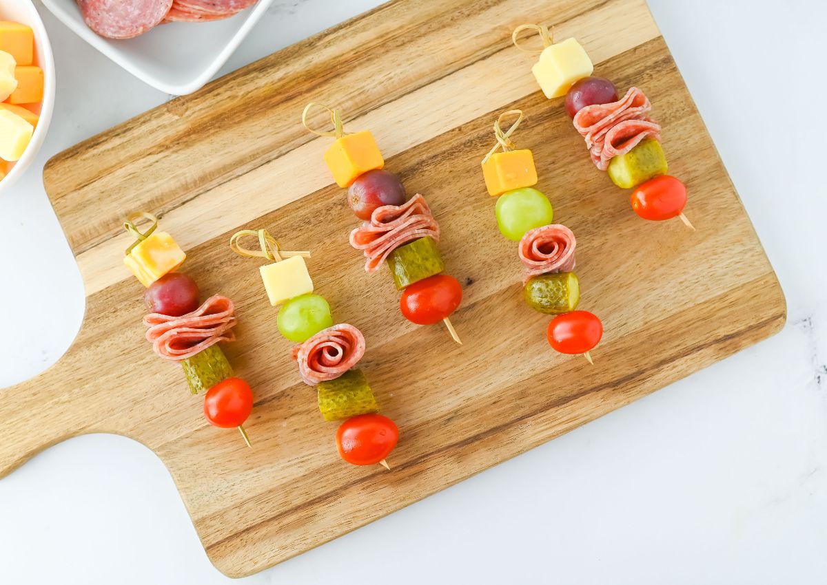 Four appetizer skewers with cheese cubes, salami, grapes, green olives, pickles, and cherry tomatoes on a wooden cutting board.
