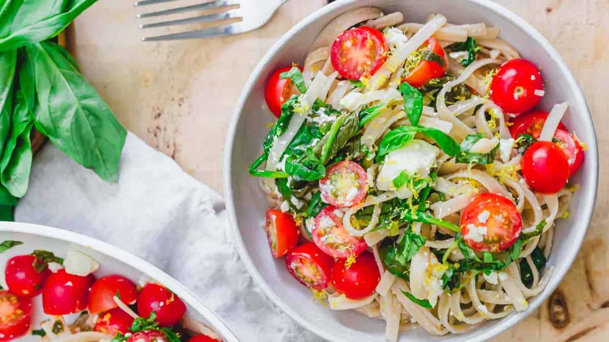 A bowl of pasta topped with cherry tomatoes, fresh basil, and grated cheese sits on a table next to a fork and fresh basil leaves.