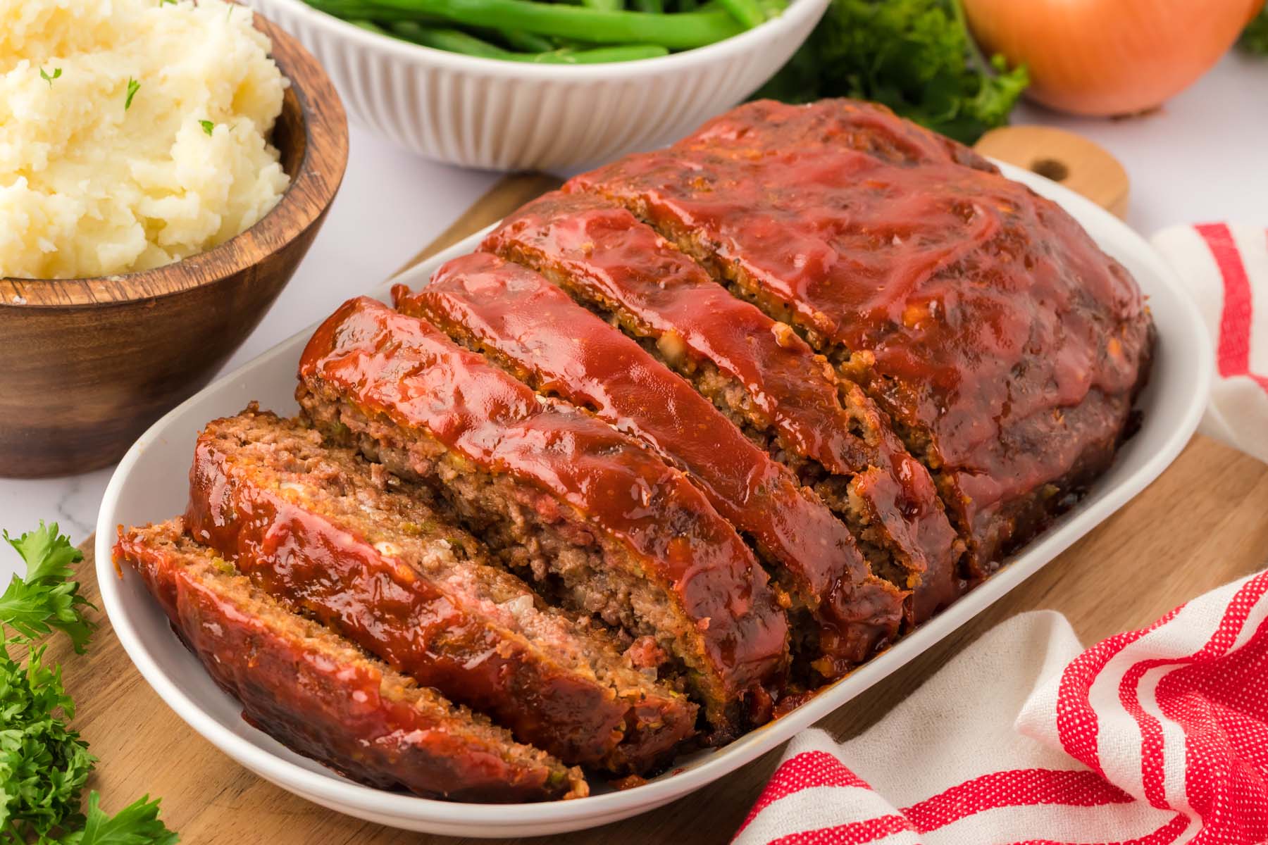 A sliced meatloaf with a ketchup glaze on a white dish, served alongside a bowl of mashed potatoes and a bowl of green beans.