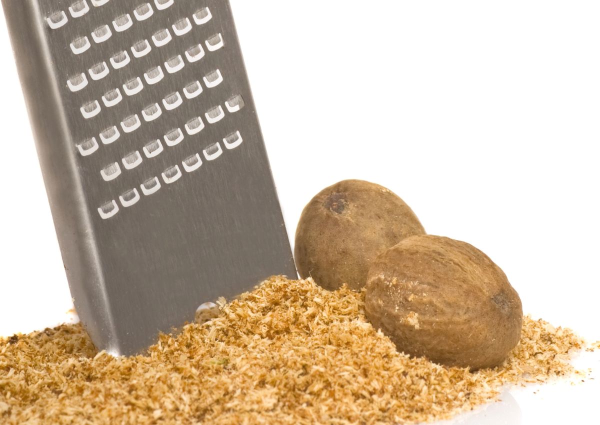 A metal grater with grated nutmeg and two whole nutmeg seeds on a white surface.
