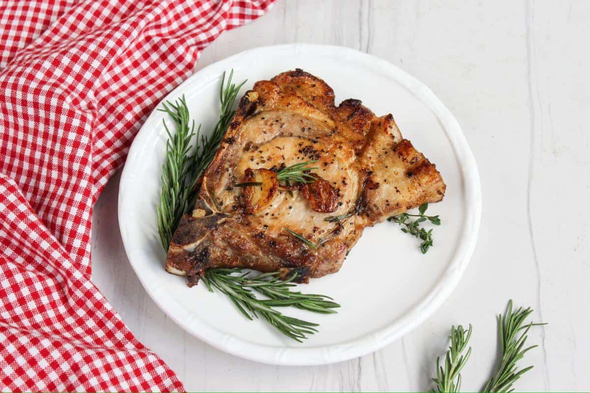 Grilled pork chop garnished with fresh rosemary and thyme on a white plate next to a red and white checkered cloth on a white surface.