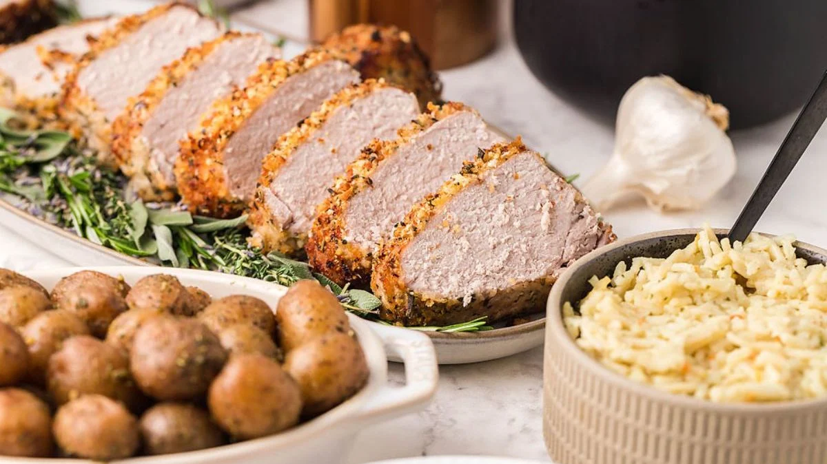 Sliced roasted pork loin on a platter with herbs, served alongside seasoned baby potatoes and a bowl of rice.