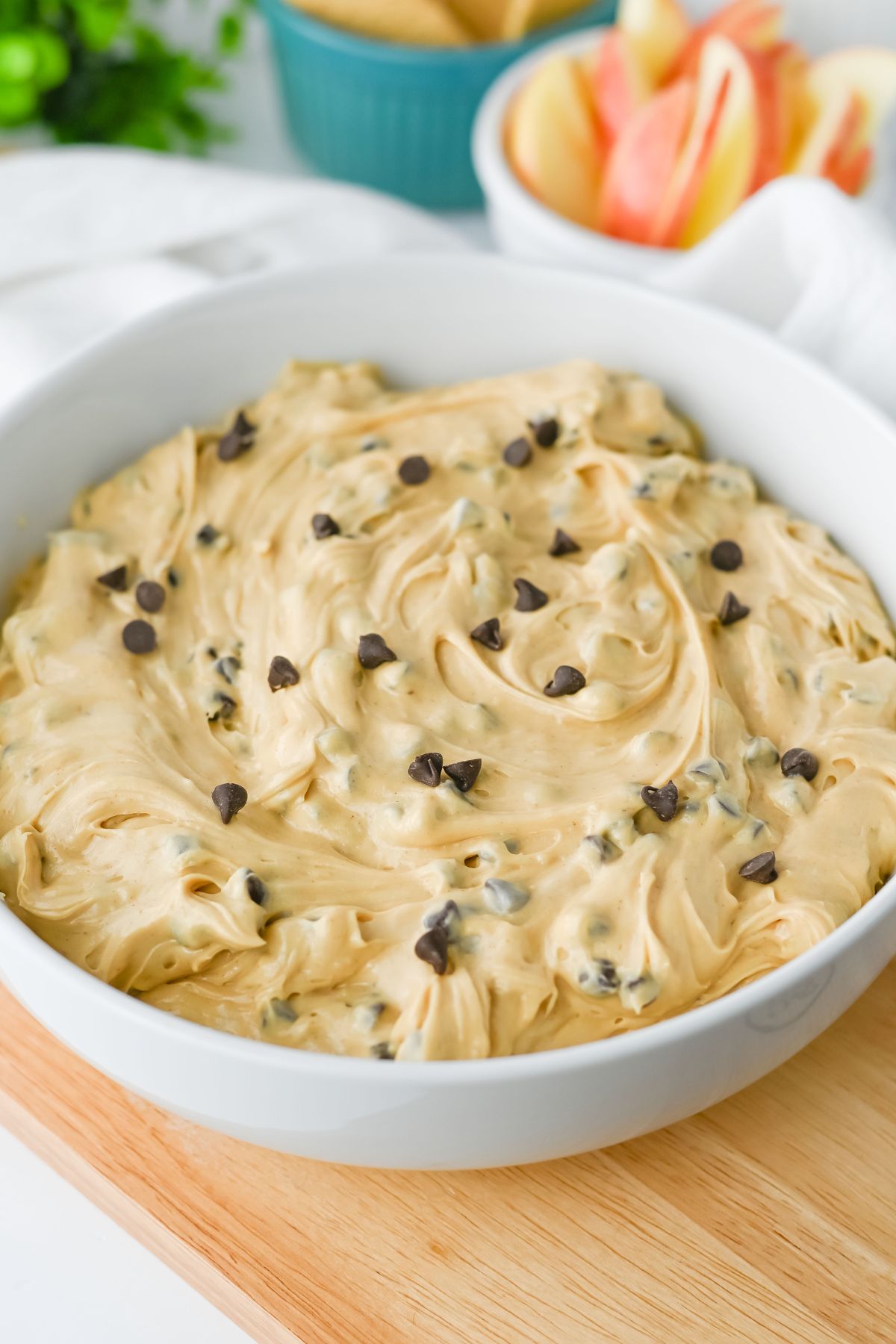 A white bowl filled with creamy dip topped with chocolate chips, placed on a wooden board with sliced apples and tortilla chips in the background.
