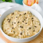 A white bowl filled with creamy cookie dough dip topped with chocolate chips, placed on a wooden surface with apple slices in the background.